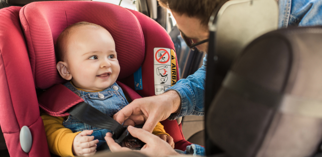 Smiling baby in a red car seat, looking up as an adult fastens the seatbelt, with the car interior visible in the background