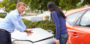 Two people appear to be discussing a car accident, examining the damage to the front of a silver car near an orange car