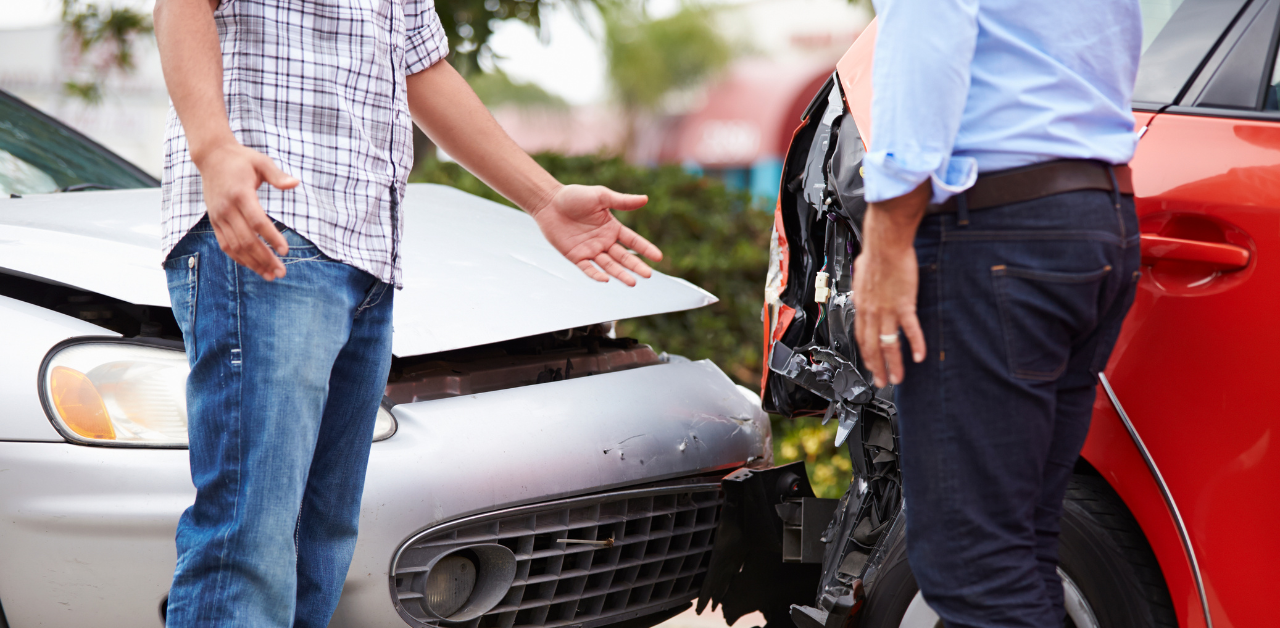 Two people standing beside their damaged cars after a collision, one gesturing with arms open, both cars visibly dented at the front