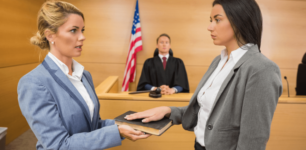 A woman places her hand on a book, taking an oath while another woman holds the book in a courtroom, with a judge observing in the background