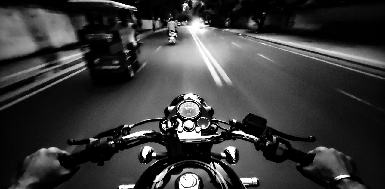 A black-and-white view from a motorcycle rider's perspective on a city street at night, showing handlebars and distant vehicles