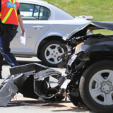 A black car with severe front-end damage in a collision, with a police officer in a reflective vest inspecting the scene nearby
