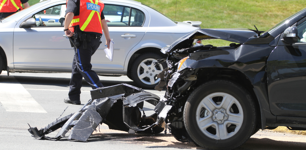 A black car with severe front-end damage in a collision, with a police officer in a reflective vest inspecting the scene nearby