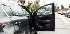 A black car with the driver's door open, parked on a residential street with other cars in the background on a rainy day