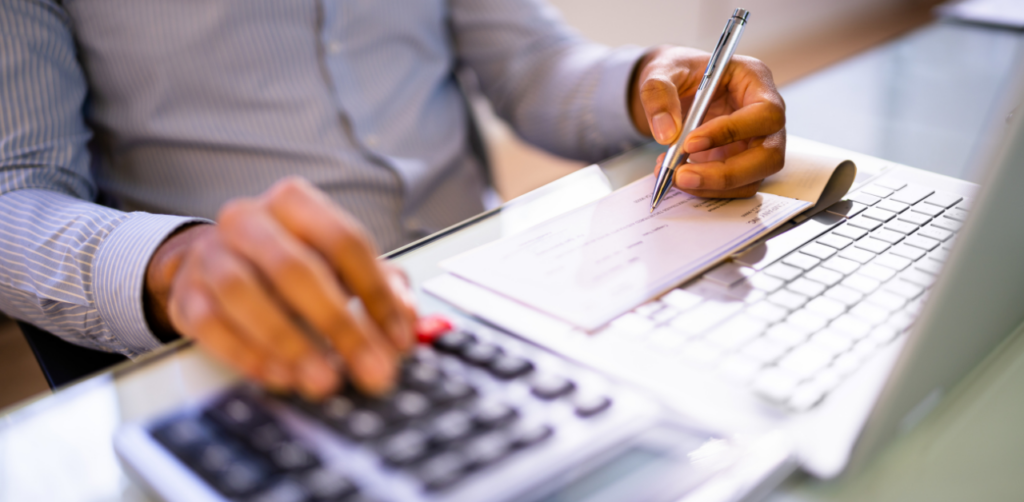 A close-up of a person writing a check while using a calculator and a laptop on a desk