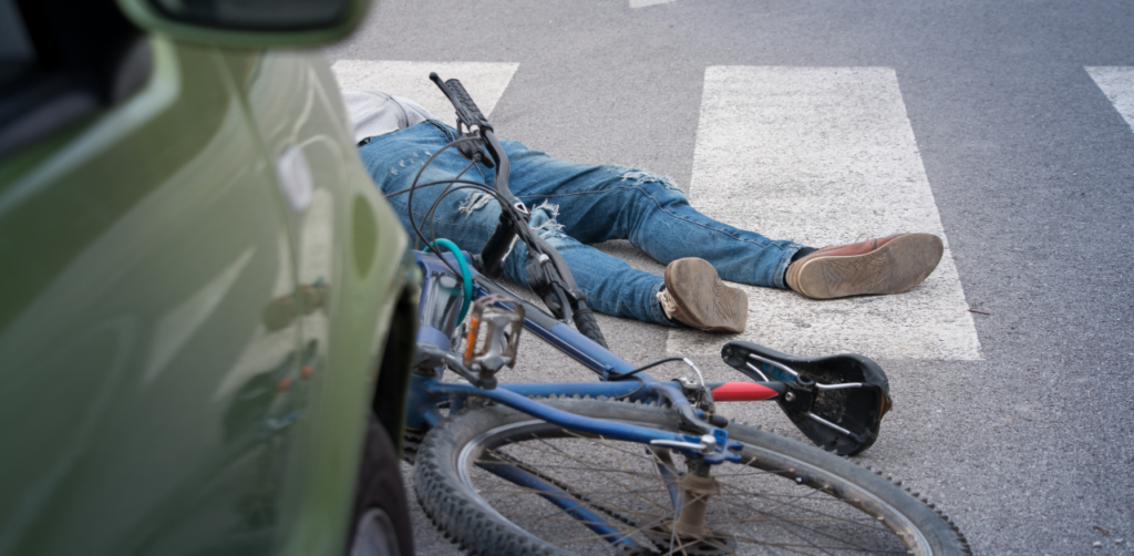 A cyclist lying on the ground near a green car at a pedestrian crossing, with a bicycle overturned beside them