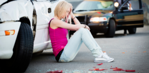 A distressed young woman sitting on the ground next to a damaged white car after an accident, with broken glass and debris around her