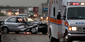 A heavily damaged car on a rainy highway with an ambulance nearby and traffic in the background