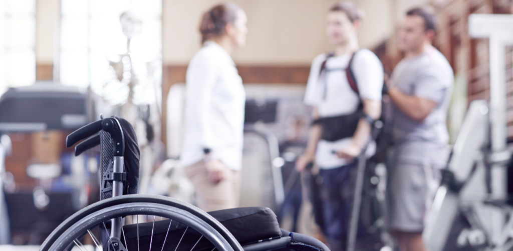 A wheelchair in the foreground with three people in the background, one wearing a harness, in a physical therapy or rehabilitation setting