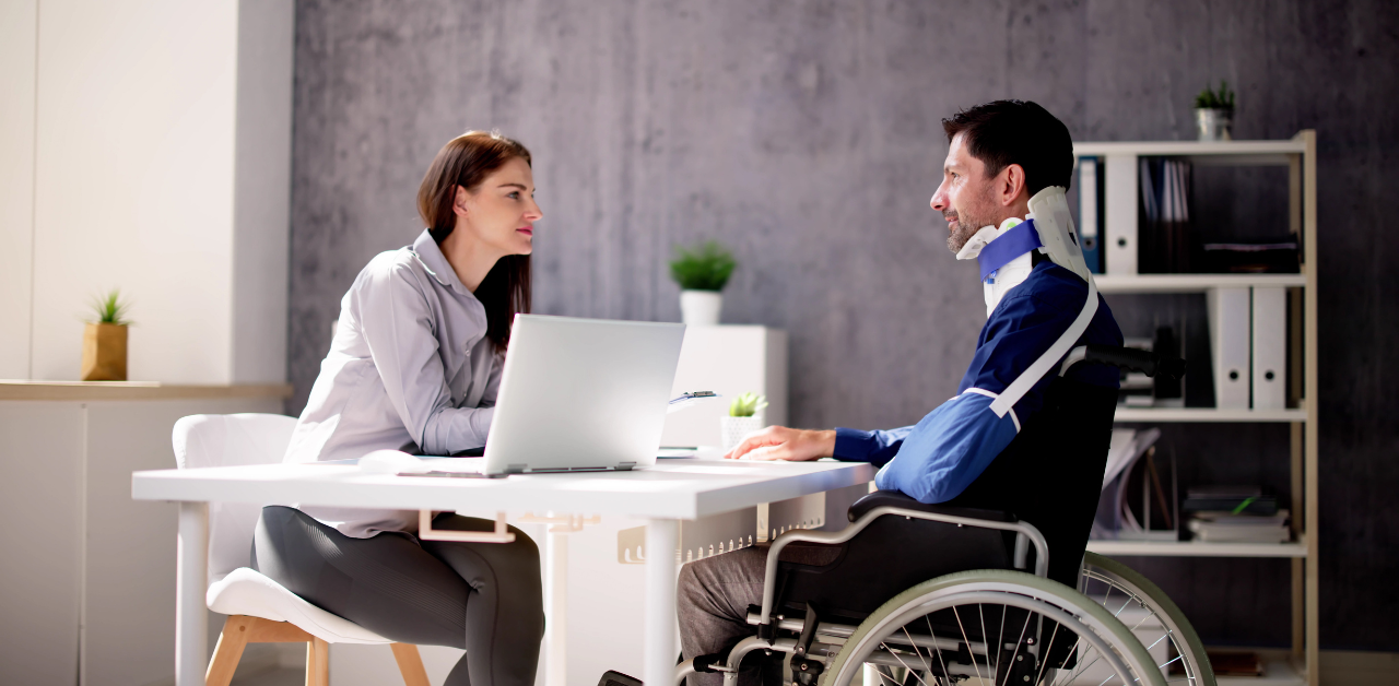 A woman in business attire talks to a man in a wheelchair with a neck brace, sitting across a desk in an office setting