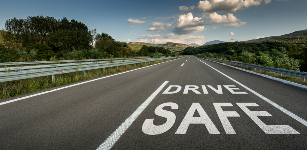 An empty highway with Drive Safe painted on the road, surrounded by greenery and mountains under a partly cloudy sky