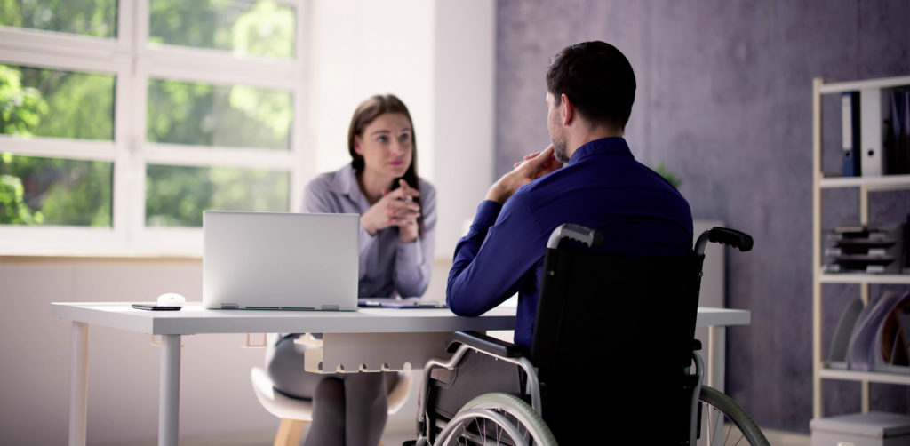 Man in a wheelchair speaking with a woman at a desk, both engaged in conversation in an office setting