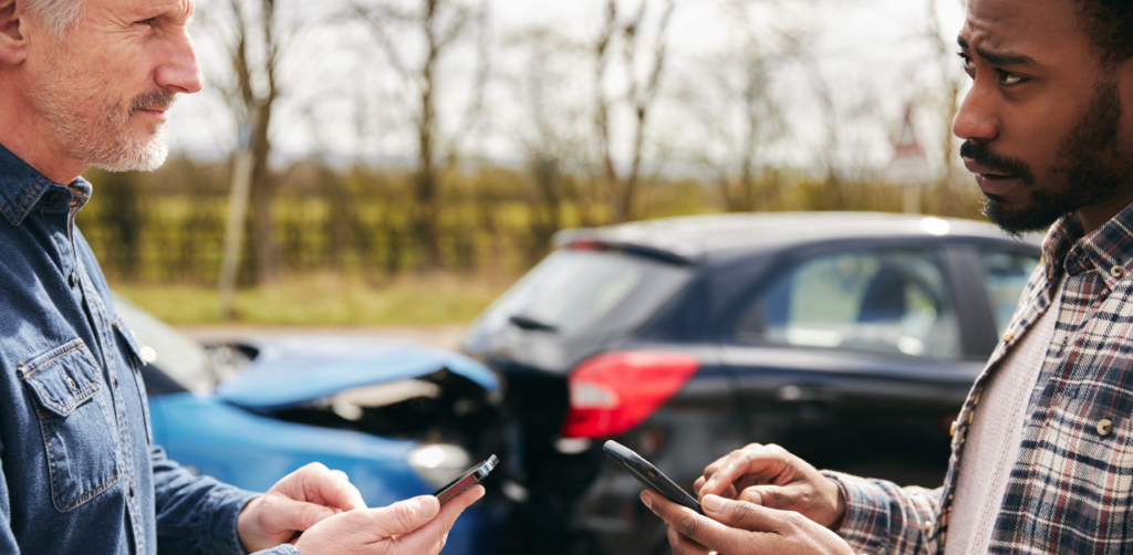 Two men exchanging insurance information on their phones after a car accident, with damaged vehicles in the background