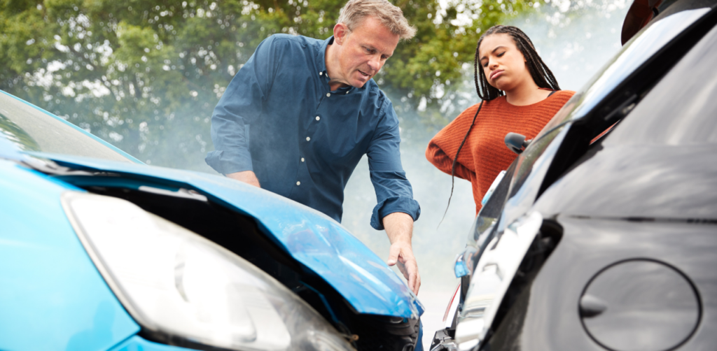 Two people examining damage to the front of two cars after a collision, with smoke rising in the background