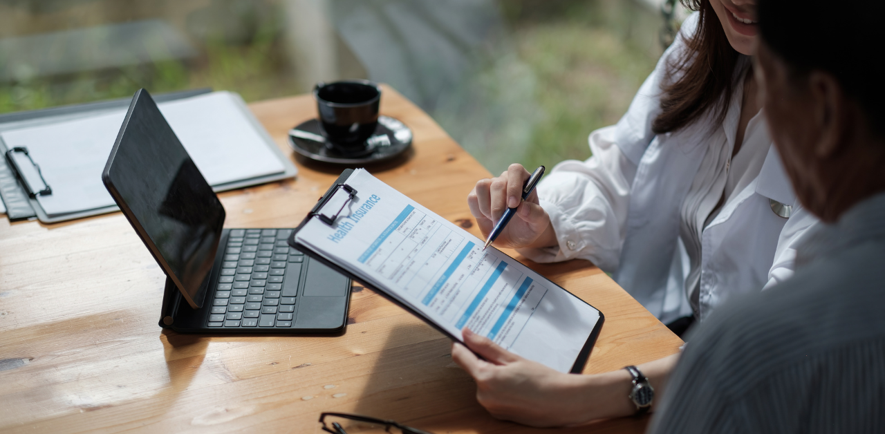 A doctor in a white coat explaining a health insurance form to a patient at a table with a tablet and clipboard