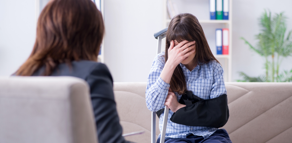 A woman with her arm in a sling sitting on a couch, covering her face while talking to another person