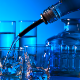 Close-up of vodka or clear liquid being poured into a shot glass, surrounded by multiple glasses and a decorative glass bottle against a blue-lit background