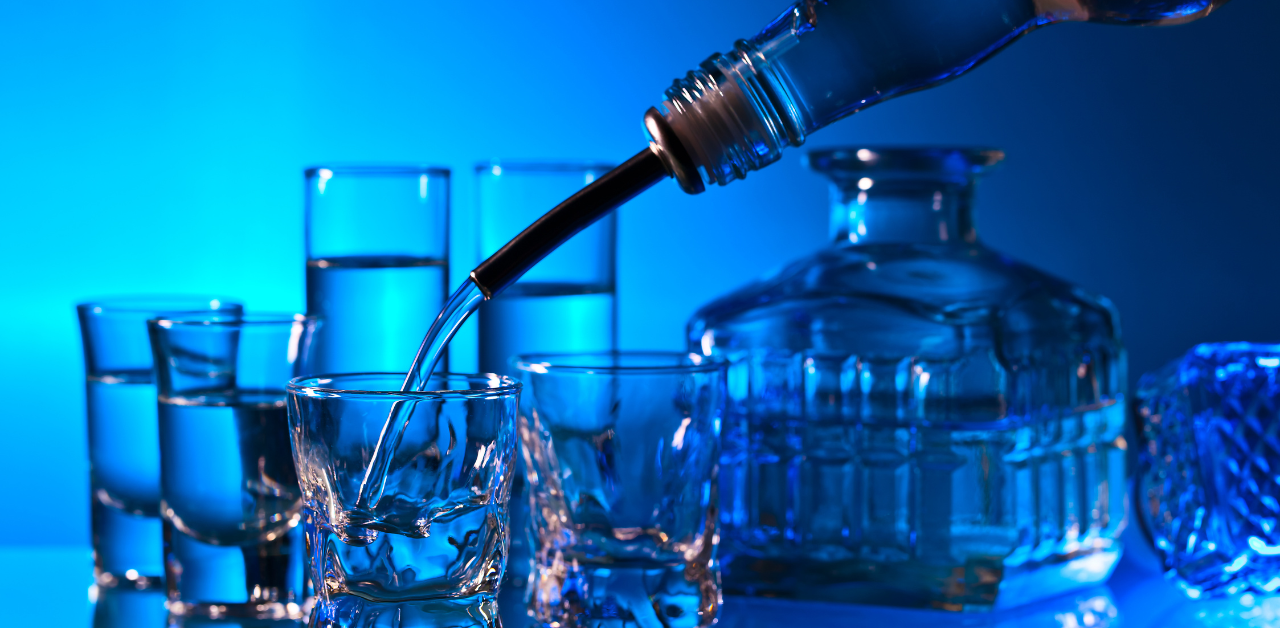 Close-up of vodka or clear liquid being poured into a shot glass, surrounded by multiple glasses and a decorative glass bottle against a blue-lit background