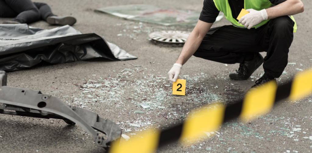 Crime scene investigator in gloves and a reflective vest examines shattered glass and car debris on a road, marking evidence with a yellow 2 marker