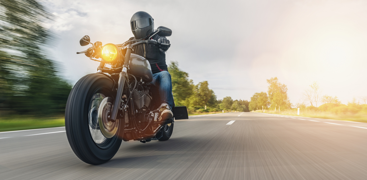 Motorcyclist wearing a helmet and black gear rides a motorcycle on an open road surrounded by trees, with sunlight in the background