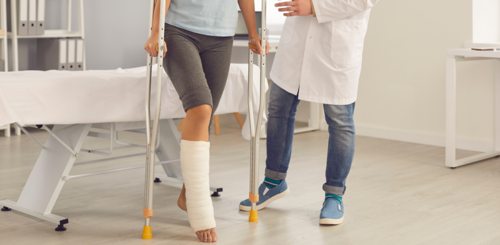 Person with a cast on their lower leg using crutches in a medical office, assisted by a doctor