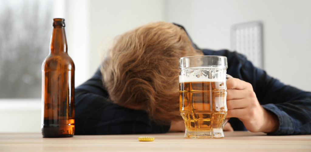 Person with their head down on a table holds a glass of beer, with an empty beer bottle and cap nearby