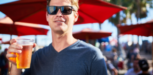 Smiling man wearing sunglasses and a gray t-shirt holds a cup of beer while standing in front of red umbrellas at an outdoor event