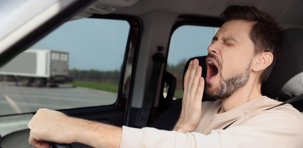 Tired man yawning while driving, one hand on the steering wheel, suggesting driver fatigue
