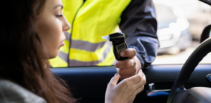Woman inside a car holds a breathalyzer device handed to her by a person wearing a reflective yellow vest, likely during a roadside check
