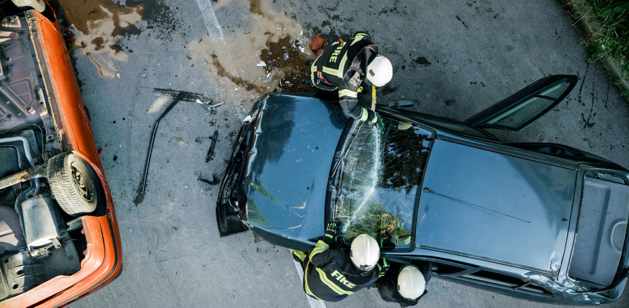 Aerial view of firefighters rescuing a person from a crashed black car near an overturned orange vehicle, with debris scattered on the road