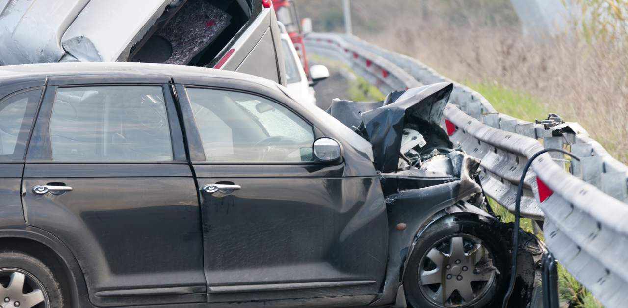 Black car with severe front-end damage crashed into a guardrail, with emergency vehicles in the background