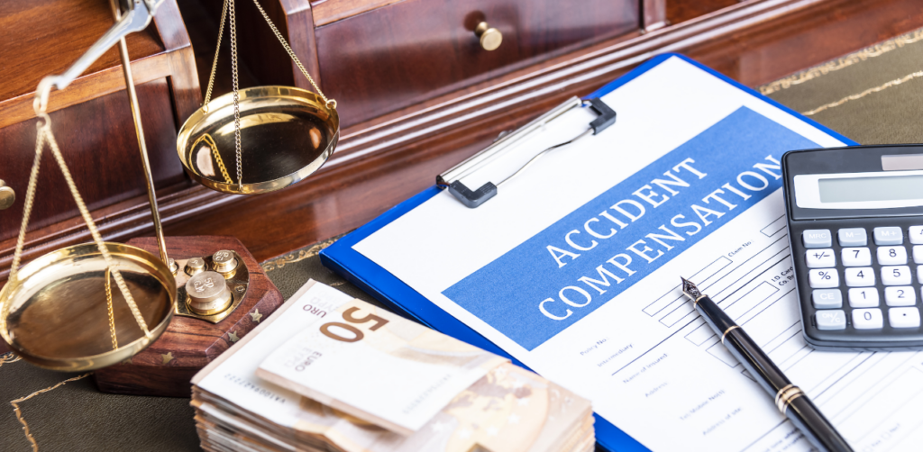 Clipboard with an Accident Compensation form, a calculator, a pen, stacks of euro banknotes, and golden scales of justice on a wooden desk