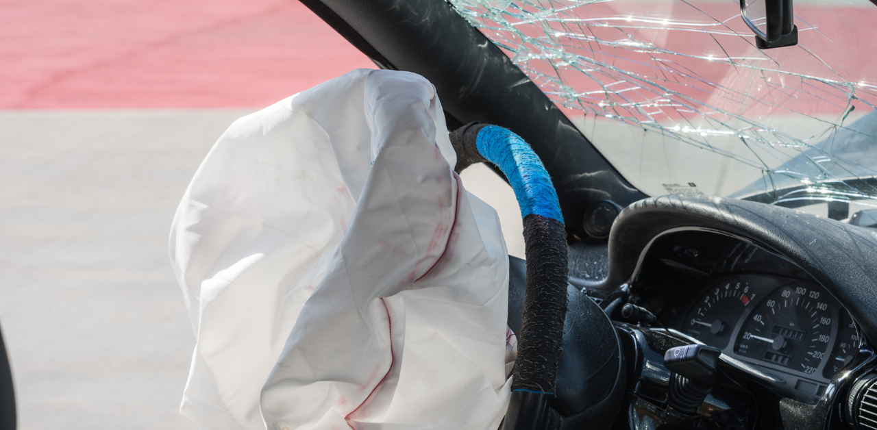 Deployed airbag with blood stains inside a car with a shattered windshield and a steering wheel covered in blue and black tape