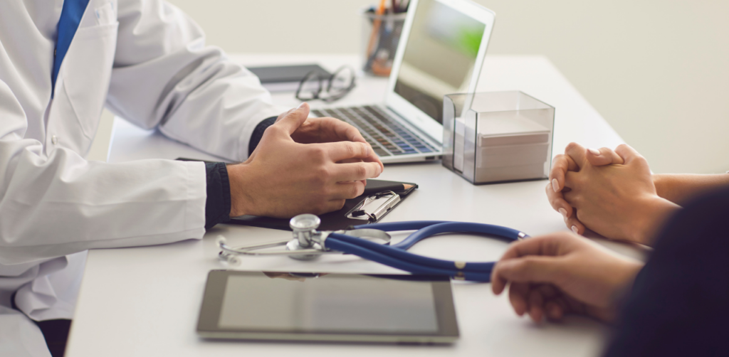 Doctor in a white coat talks with a patient at a desk, with a stethoscope, tablet, and laptop in the foreground