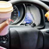 Driver holds a coffee cup in one hand and a sandwich in the other while gripping the steering wheel, illustrating distracted driving