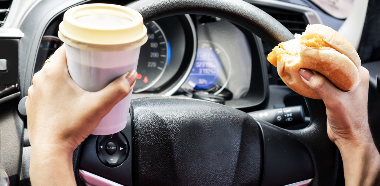 Driver holds a coffee cup in one hand and a sandwich in the other while gripping the steering wheel, illustrating distracted driving