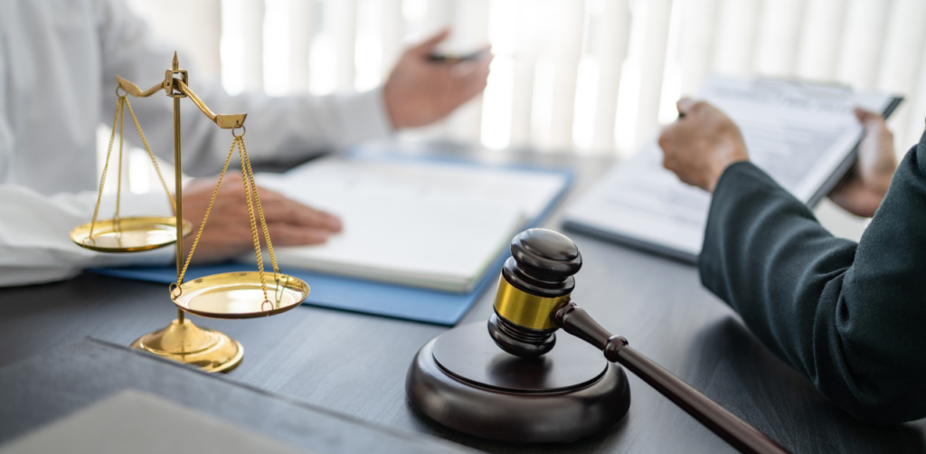 Gavel and scales of justice on a desk, with two people in suits discussing legal documents in the background