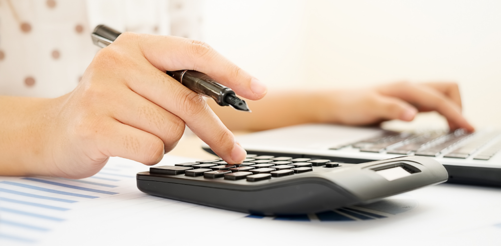 Hand pressing a calculator while using a laptop, with documents and charts in the background