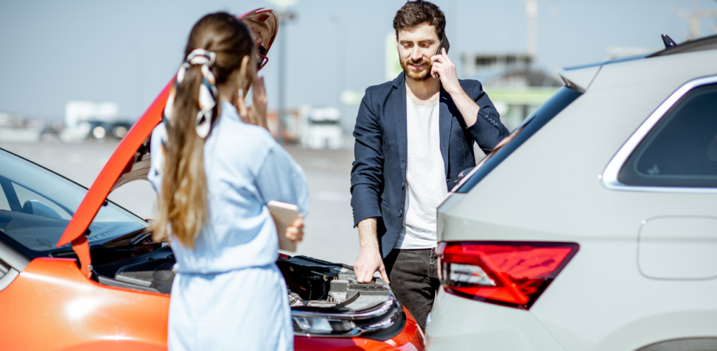 Man and a woman stand near two cars involved in a minor accident. The man is on the phone, while the woman holds a smartphone near a red car with an open hood