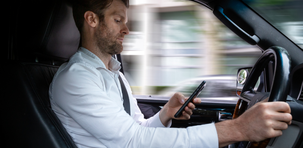 Man in a white shirt is driving while looking at his phone, with a blurred cityscape visible through the window, suggesting movement