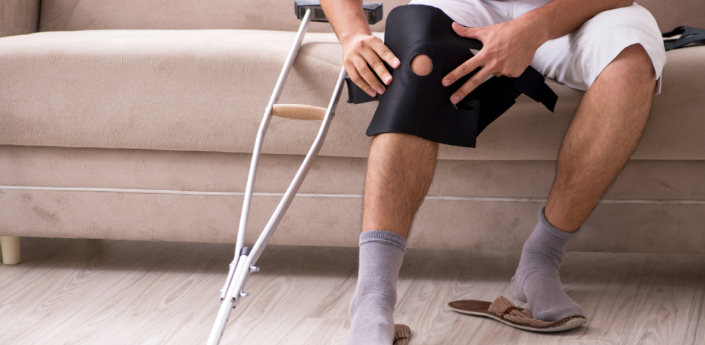 Man sitting on a couch puts on a knee brace, with crutches beside him, indicating recovery from an injury
