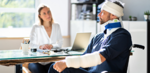 Man with head and neck bandages, wearing a neck brace, sits in a wheelchair while talking to a woman at a desk with a laptop and papers