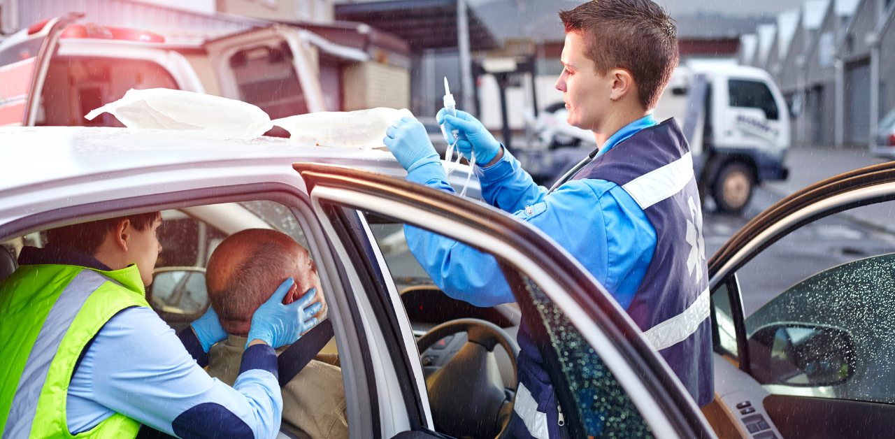 Paramedics assist an injured man inside a car, with one holding his head and another preparing a syringe, while an ambulance is parked nearby