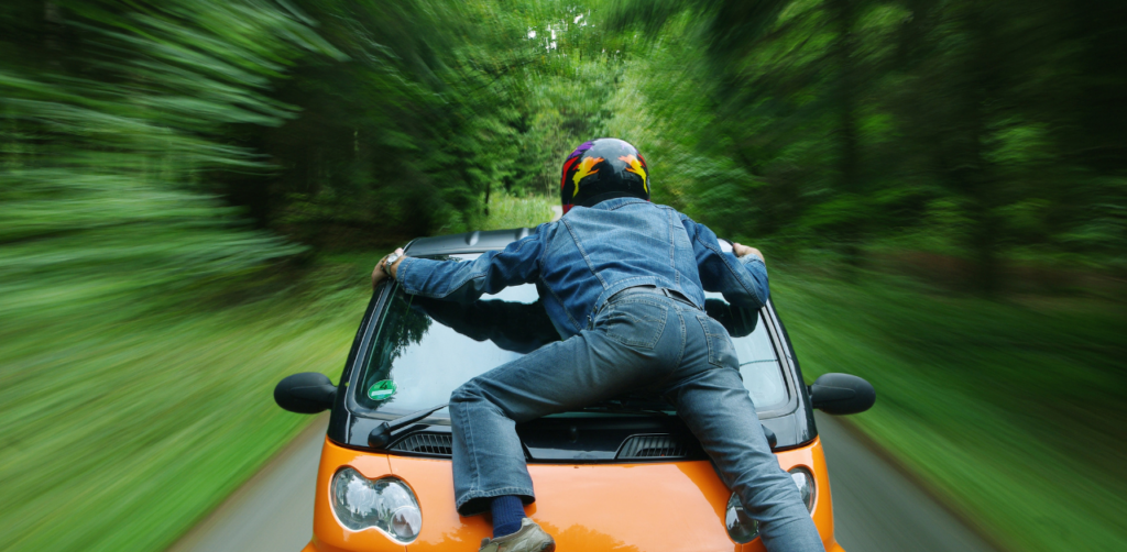 Person wearing a helmet and denim outfit clings to the hood of a moving orange car on a blurred forest road