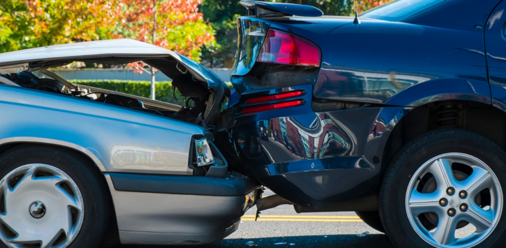 Rear-end car accident showing a silver car with a crumpled hood that collided with the back of a dark blue car on a sunny day