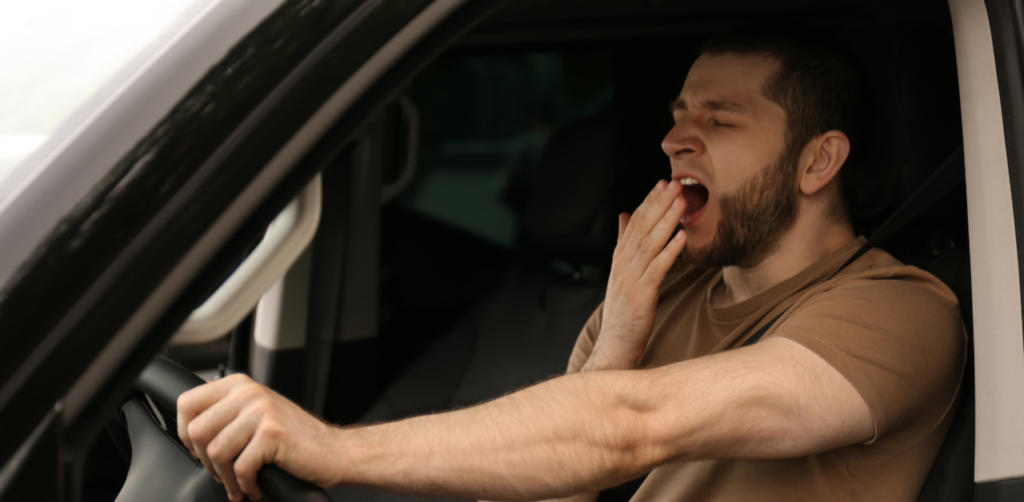 Tired man yawns while driving, with one hand on the steering wheel and the other covering his mouth