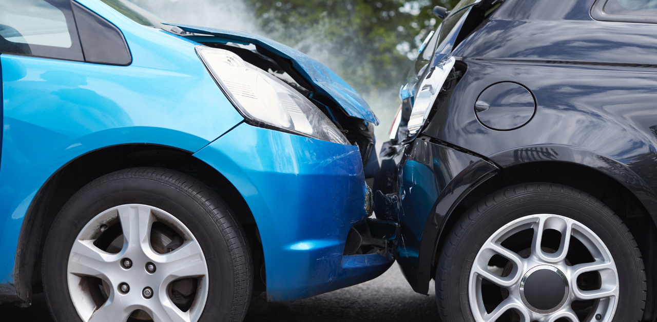 Two cars, a blue and a black one, have collided, with the blue car's hood crumpled against the black car’s rear. Smoke is visible in the background