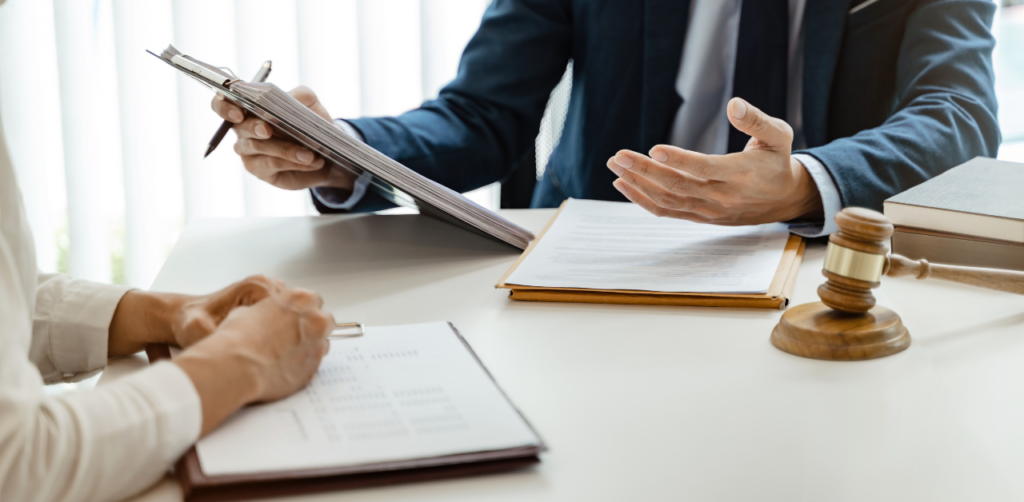 Two people seated at a desk with legal documents; one gestures while the other listens. A gavel and law books are visible on the desk