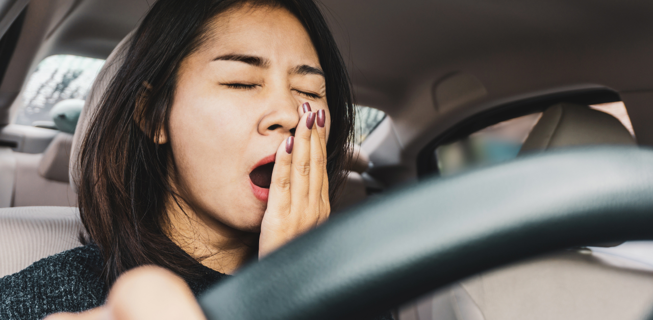 Woman sits in a car yawning with her hand covering her mouth, appearing tired while holding the steering wheel
