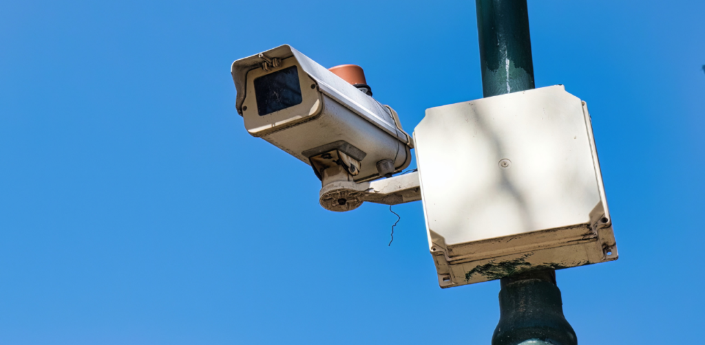 Mounted traffic surveillance camera on a metal pole against a bright blue sky, monitoring an intersection
